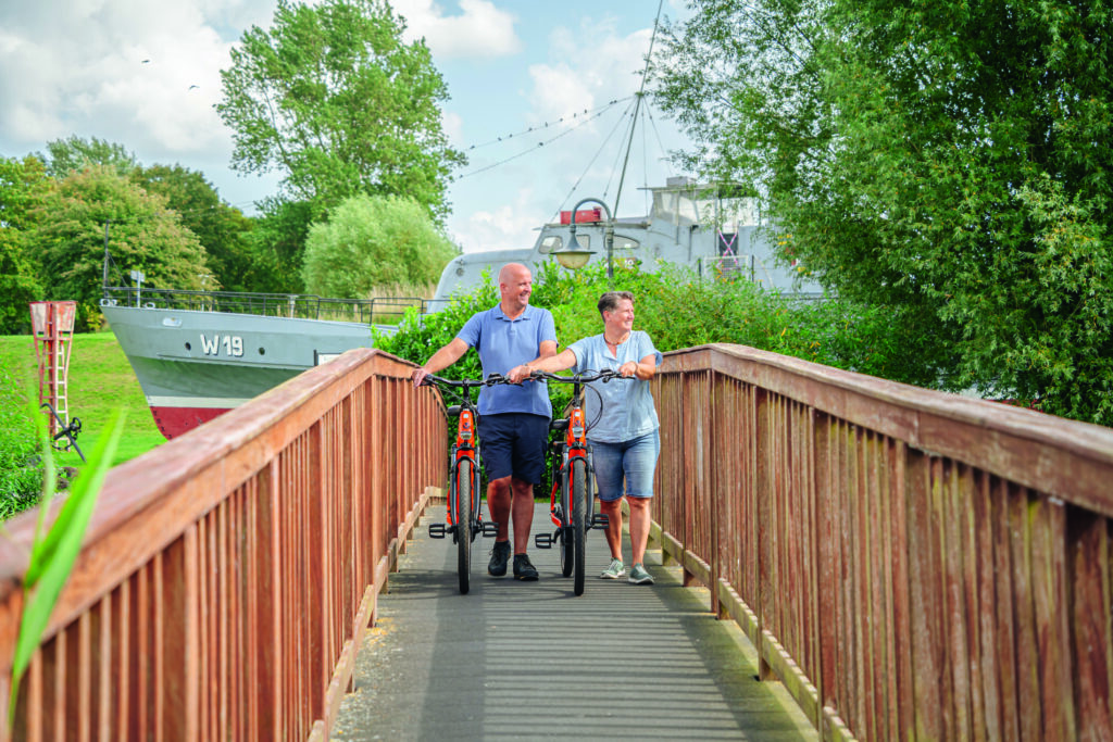 Paar schiebt Nordseeräder über Brücke mit Küstenwachboot dahinter