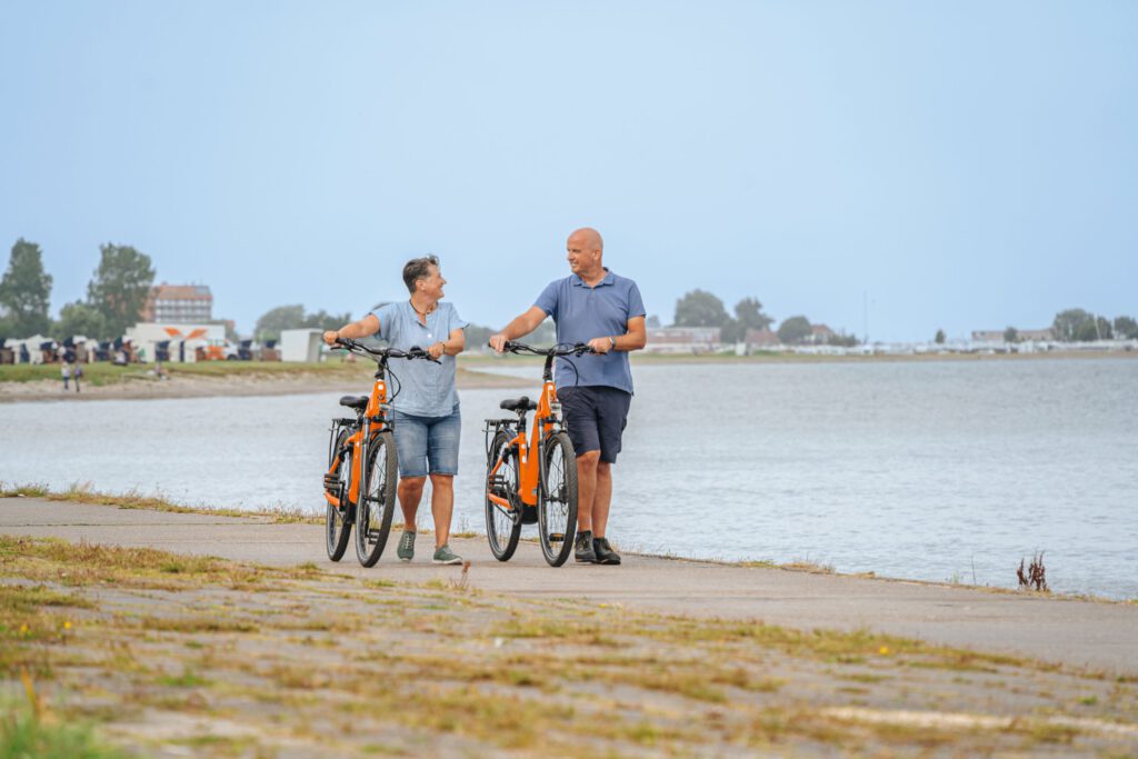 Paar schiebt Nordseeräder vor Strand/ Wasserkulisse